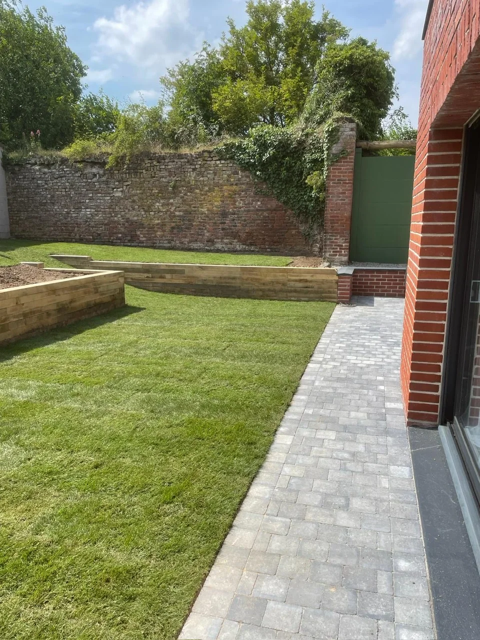 Jardin engazonné avec allée pavée, bordures en bois et mur en briques.